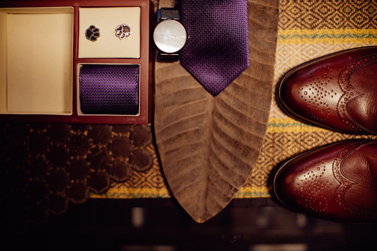 Flat lay of men's accessories featuring a purple tie, cufflinks, watch, and leather shoes.